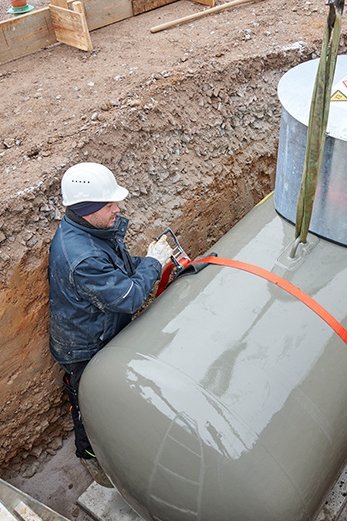 Arbeiter befestigt einen grauen Flüssiggasbehälter mit einem orangen Spanngurt in einer Baugrube