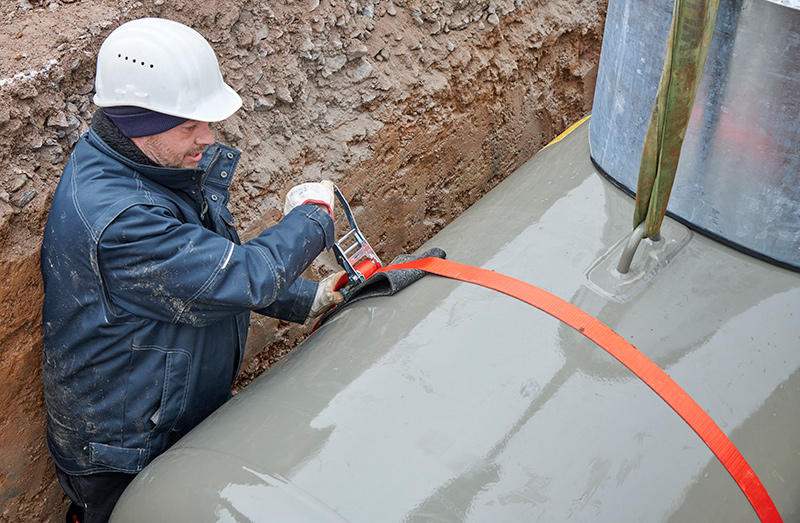 Arbeiter mit weißem Helm befestigt orangenes Spanngurtband an Flüssiggasbehälter auf Auftriebssicherung