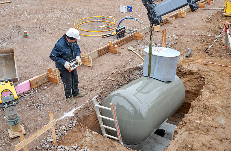 Arbeiter mit Helm setzt einen grauen Flüssiggasbehälter in eine Baugrube ein, umgeben von Baustellenmaterialien