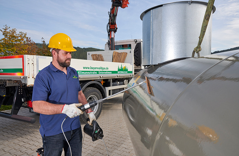 Mann mit gelbem Helm und blauer Arbeitskleidung prüft Epoxidharzbeschichtung eines großen Metallbehälters mit einem Isolationsprüfgerät vor Auslieferung