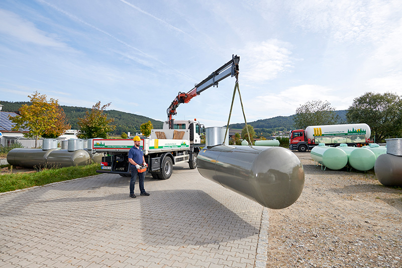 Mann mit Schutzhelm lädt großen grauen Flüssiggasbehälter mit Kran auf Lastwagen in Industrieumgebung